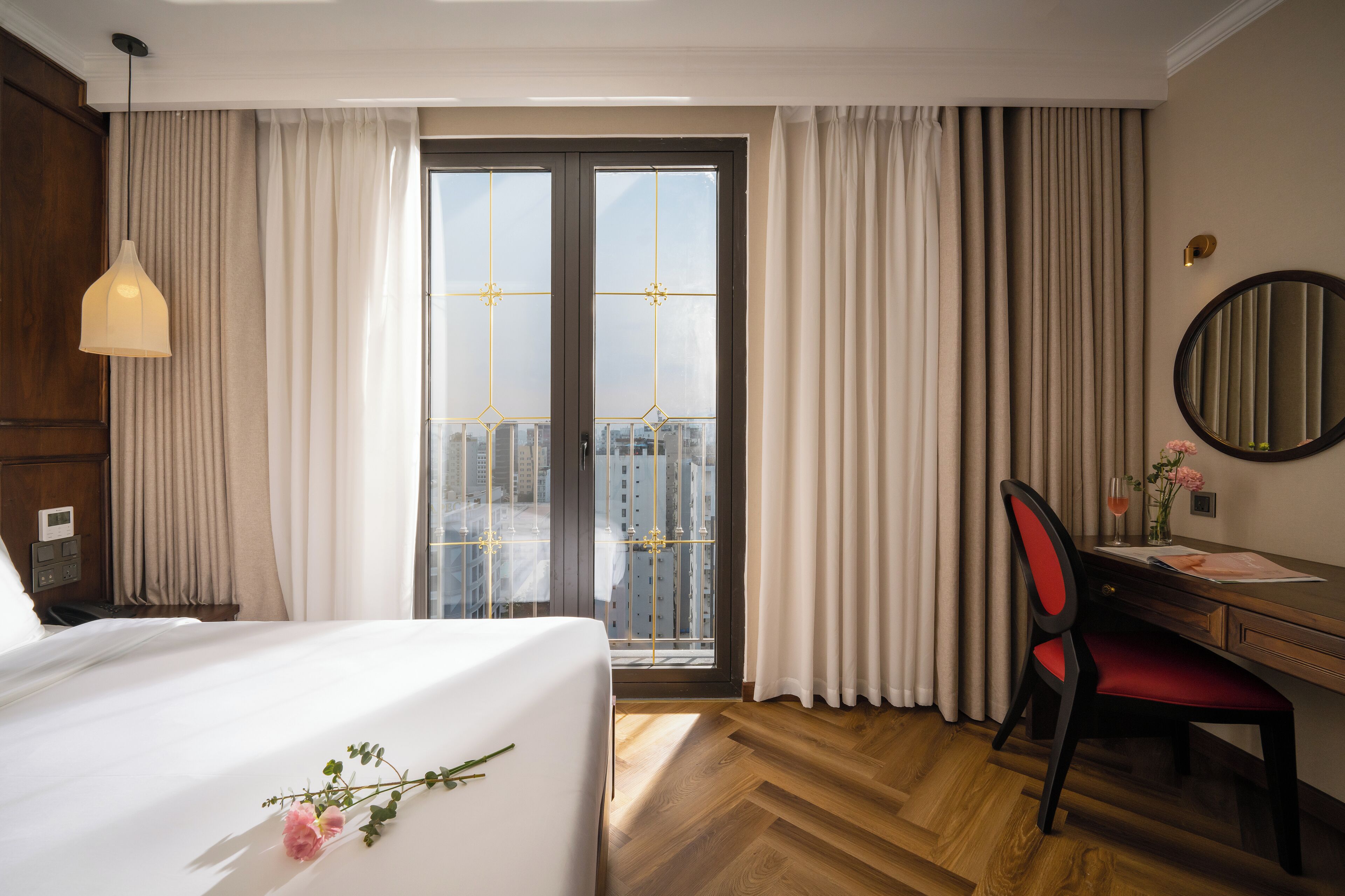 Una camera d'albergo con un letto, una scrivania, una sedia, uno specchio e vista sulla città attraverso un'ampia finestra.