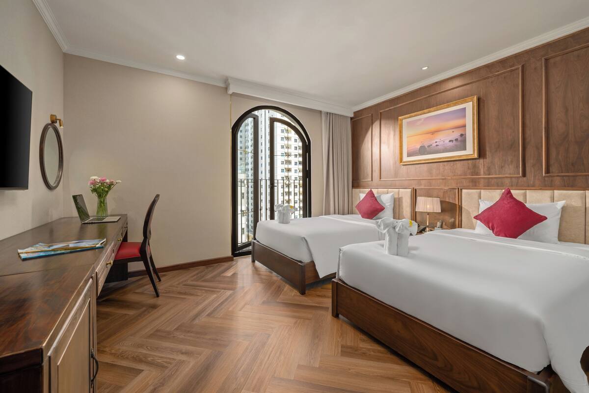 A hotel room with two beds, a desk, a chair, and a large window with a view of high-rise buildings.
