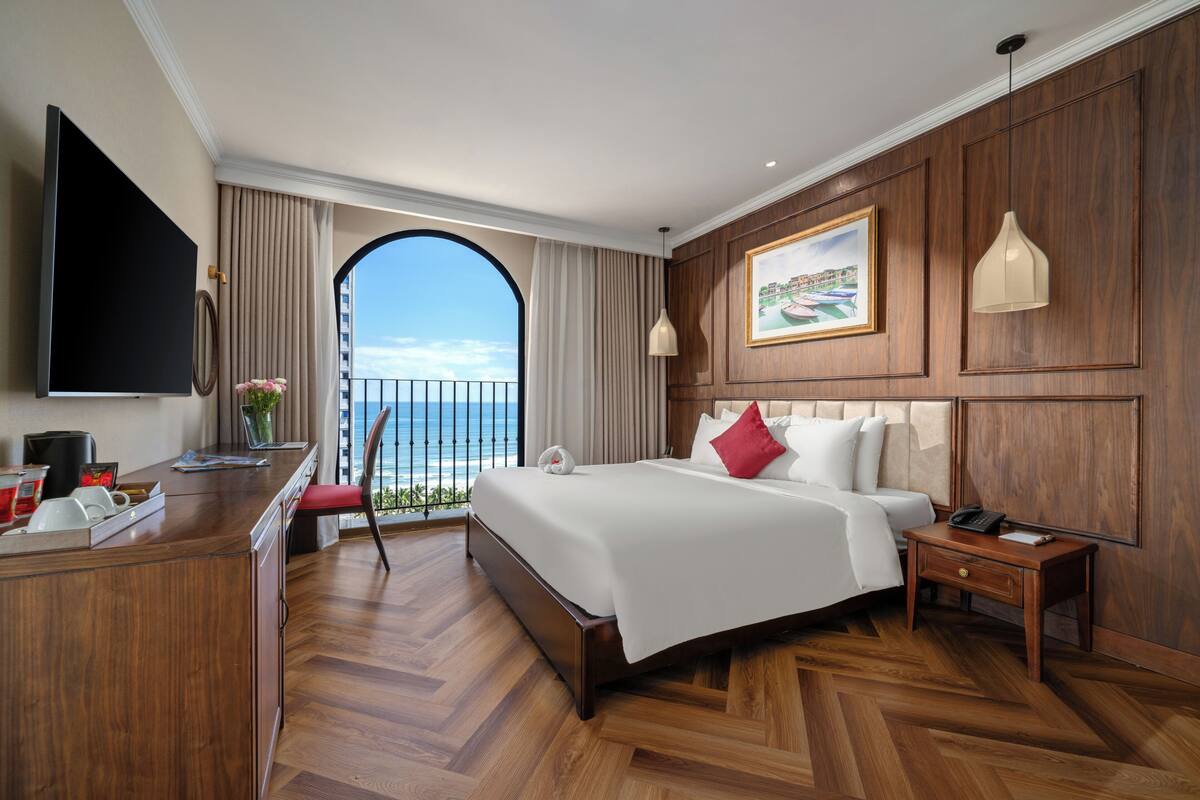 A hotel room with a large bed, a desk with a flat-screen TV, a view of the ocean through a balcony, and wooden paneling on the walls.