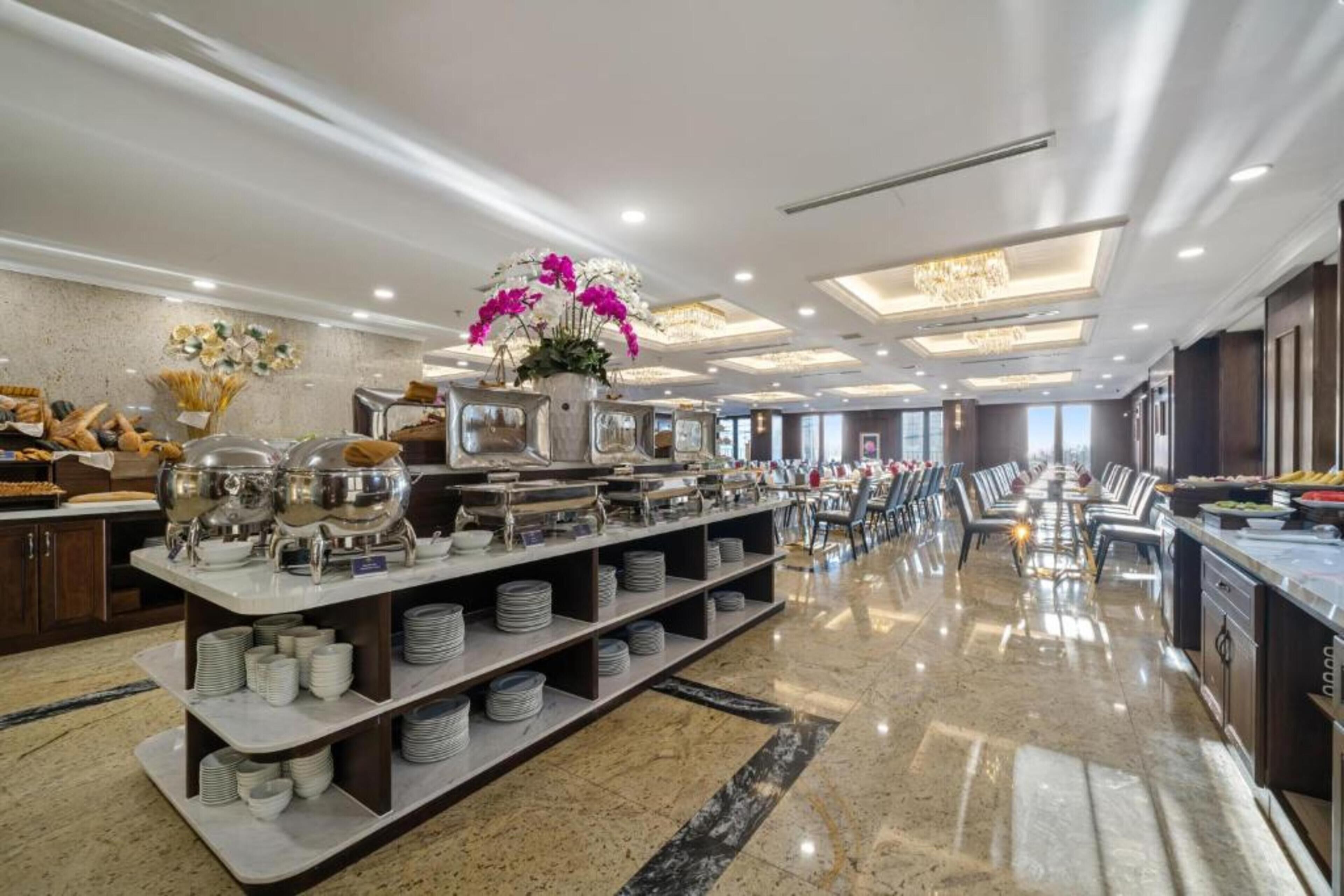 En rummelig hotelspisestue med buffet, der inkluderer flere serveringsstationer og en blomsteropsætning i midten.