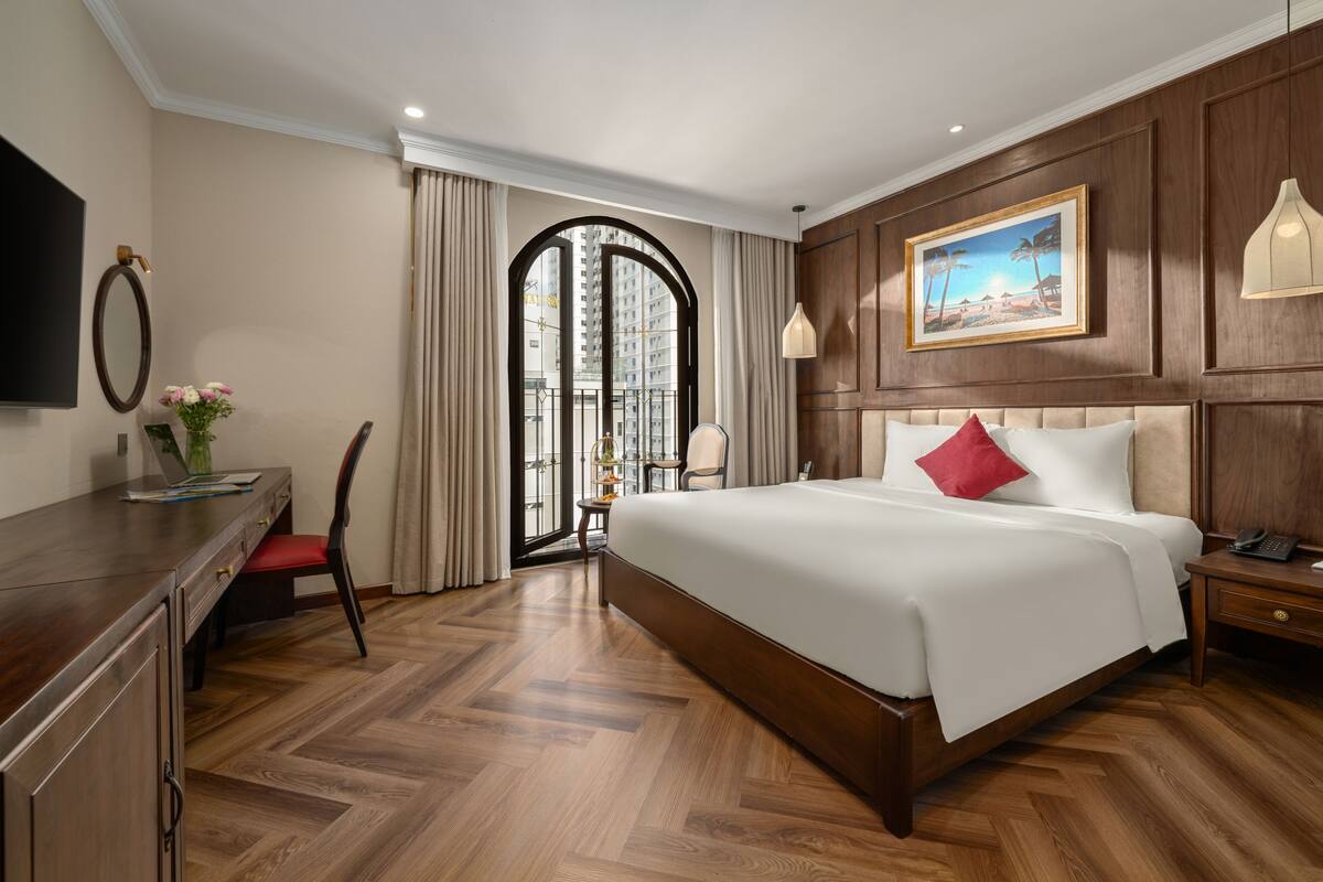 A hotel room with a large bed, a desk, a chair, a TV, a painting, and a view through a glass door.