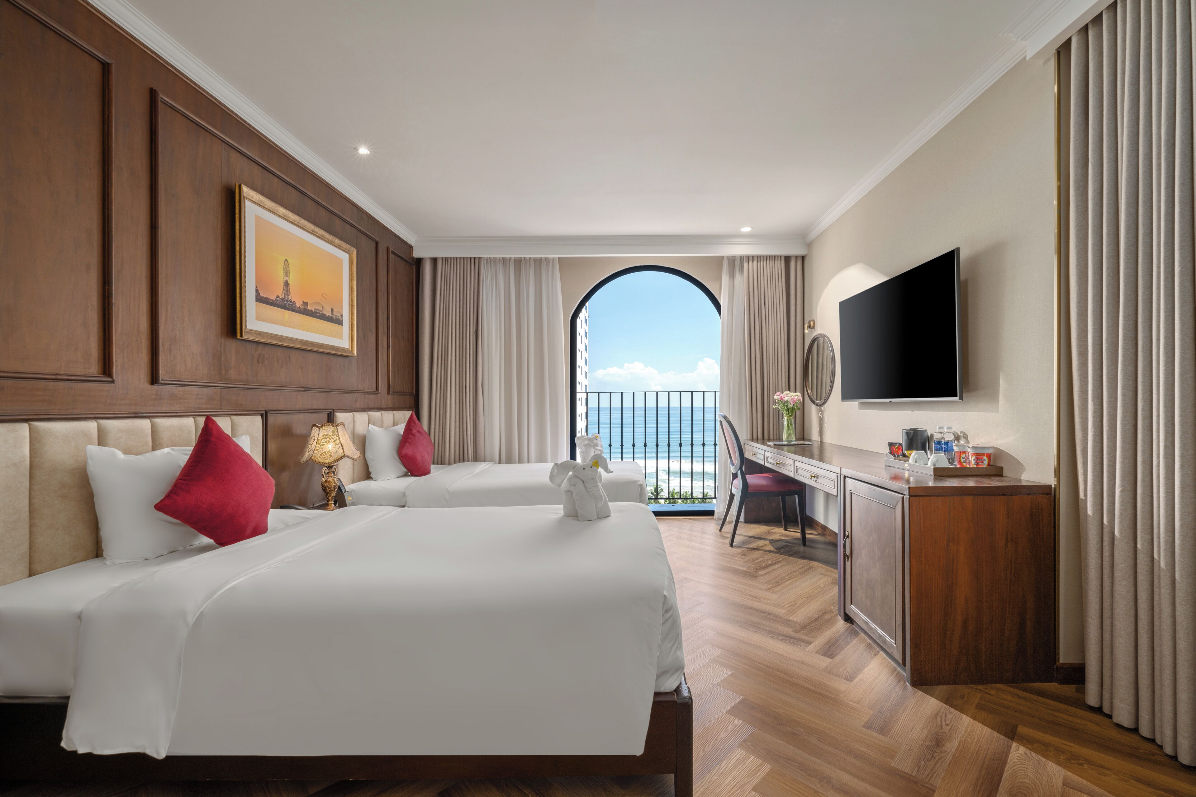 A hotel room with two beds, a desk, a TV, and a view of the sea through an arched window.