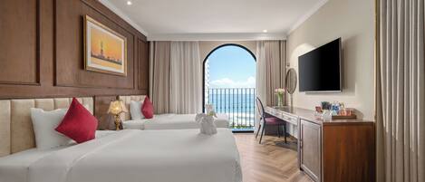 A hotel room with two beds, a desk, a TV, and a view of the sea through an arched window.