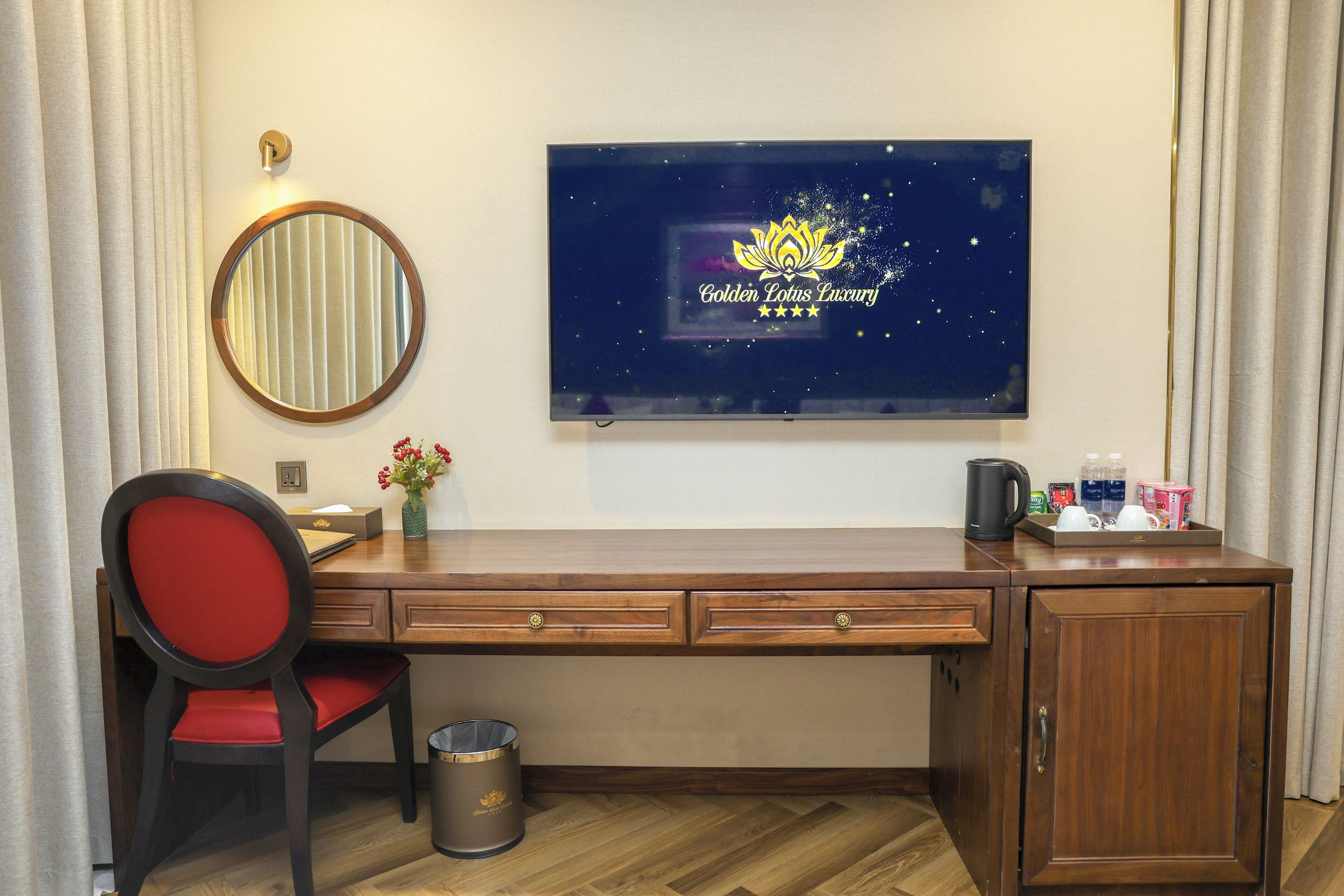 Une chambre d’hôtel avec un bureau en bois, une chaise rouge, un miroir rond, une poubelle et un téléviseur fixé au mur qui diffuse l’émission « Golden Lotus Luxury ».
