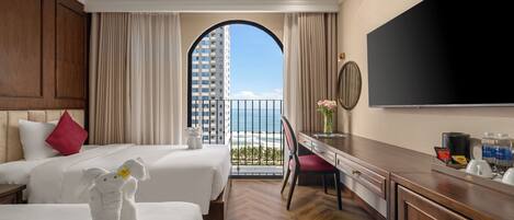 A hotel room with two beds, a desk, a TV, and a balcony with a view of the ocean.