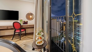 A modern hotel room with a large TV, a dining table with a tiered dessert stand, a red chair, and a city view through large windows. - Golden Lotus Luxury Hotel Da Nang (Da Nang)