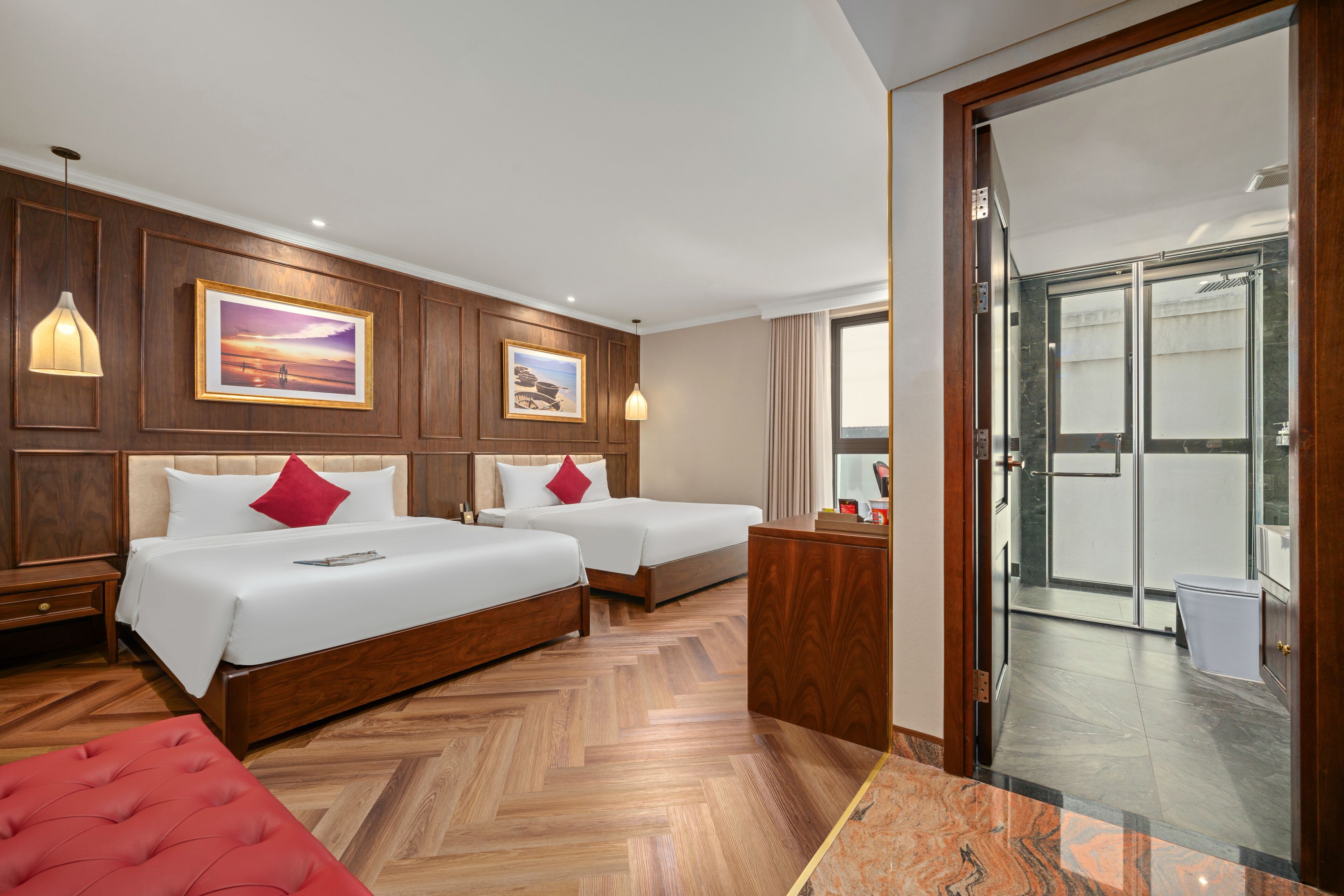 A hotel room with two beds, wooden paneling, a red ottoman, and a view of a balcony.