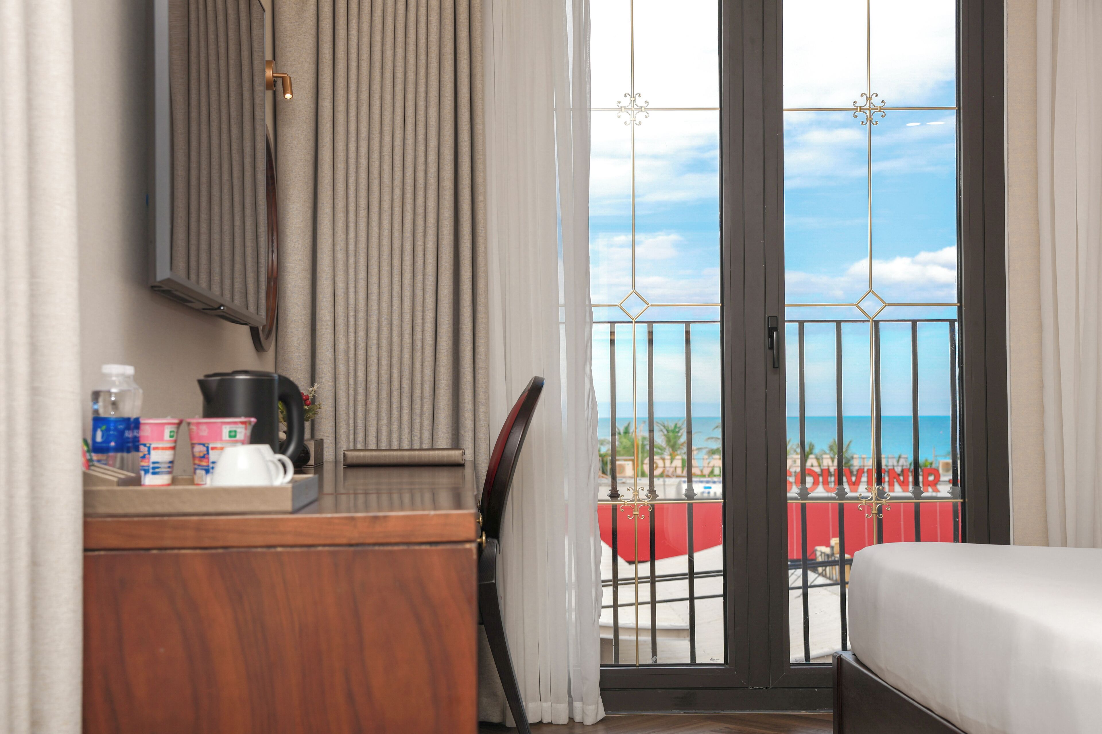 A hotel room with a balcony, a bed, a desk with toiletries, and a view of the beach.