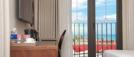 A hotel room with a balcony, a bed, a desk with toiletries, and a view of the beach.