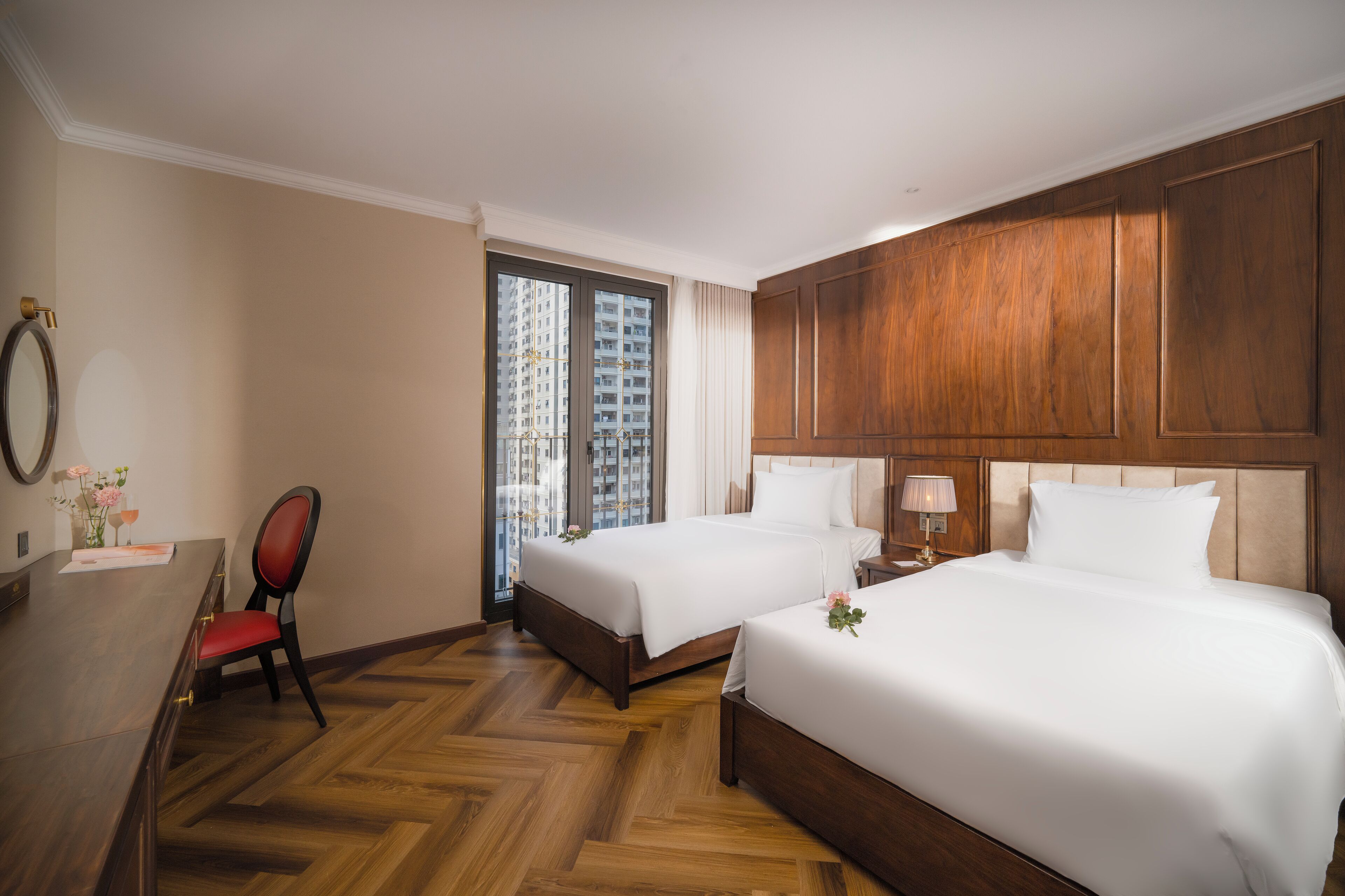 A hotel room with two beds, a desk, a chair, and a large window with a view of high-rise buildings.