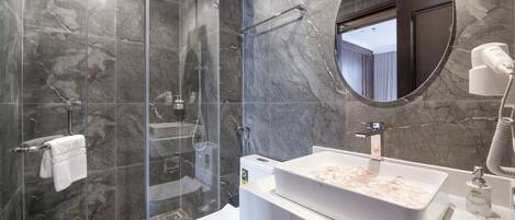Ein modernes Badezimmer mit einer begehbaren Glasdusche, einem weißen Waschbecken mit Marmorplatte und einem runden Spiegel darüber.