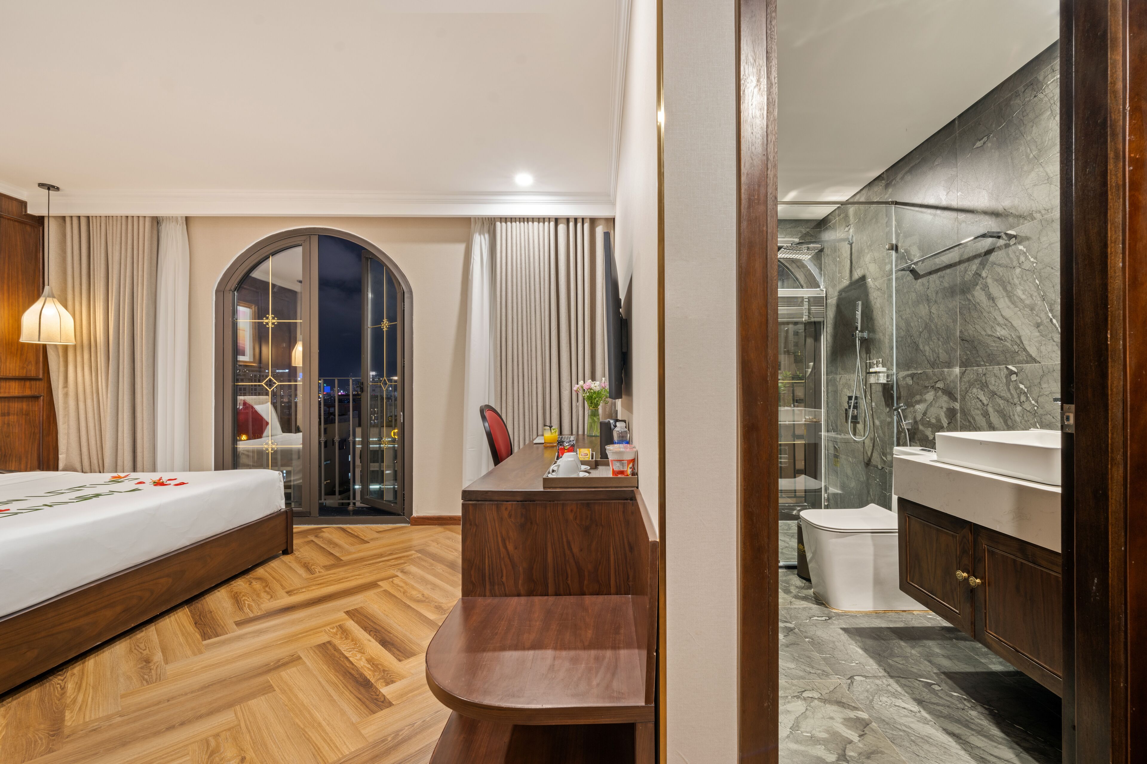 A modern hotel room with a wooden floor, a bed with a white bedspread, a wooden bench, a desk with a chair, a bathroom with a marble shower, and a large mirror.