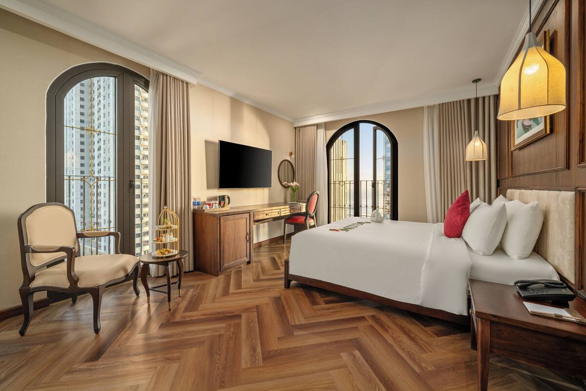 A hotel room with a large bed, a desk, a chair, a TV, and large windows.
