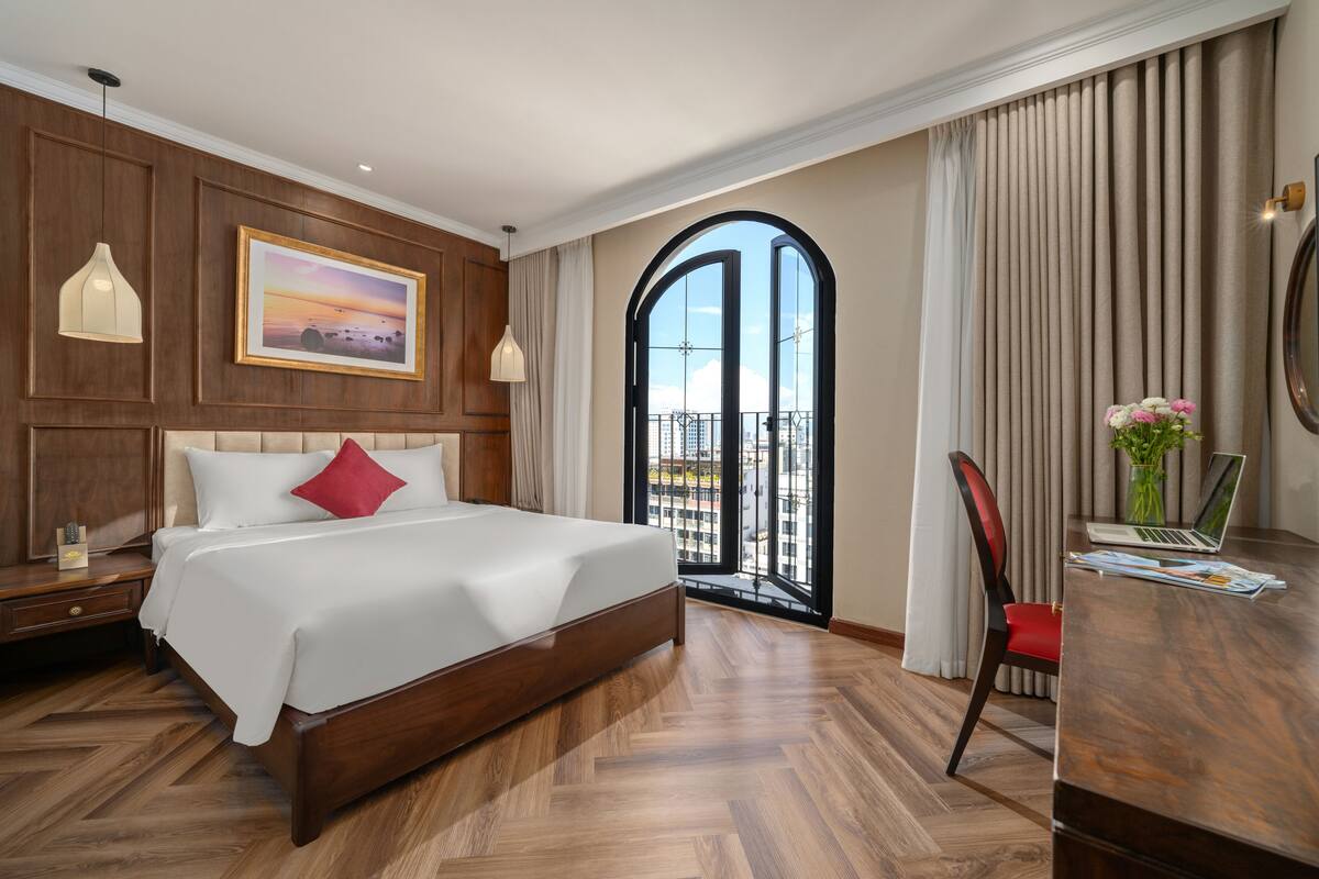 A hotel room with a large bed, a desk with a laptop, a view of the city through a large window, and a painting on the wall.