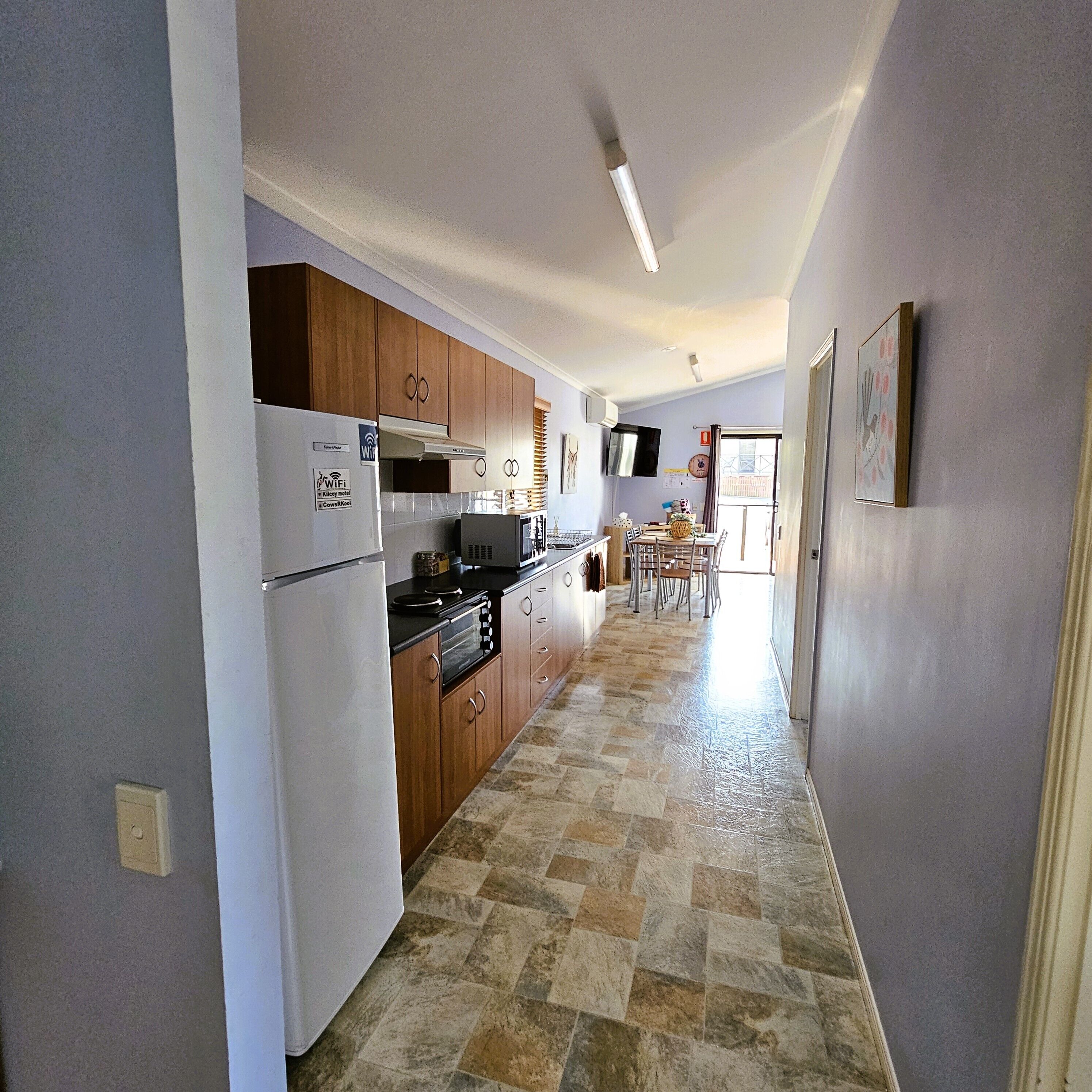 A narrow kitchen with wooden cabinets, a white refrigerator, and a microwave. A dining area with a table and chairs is visible in the background.