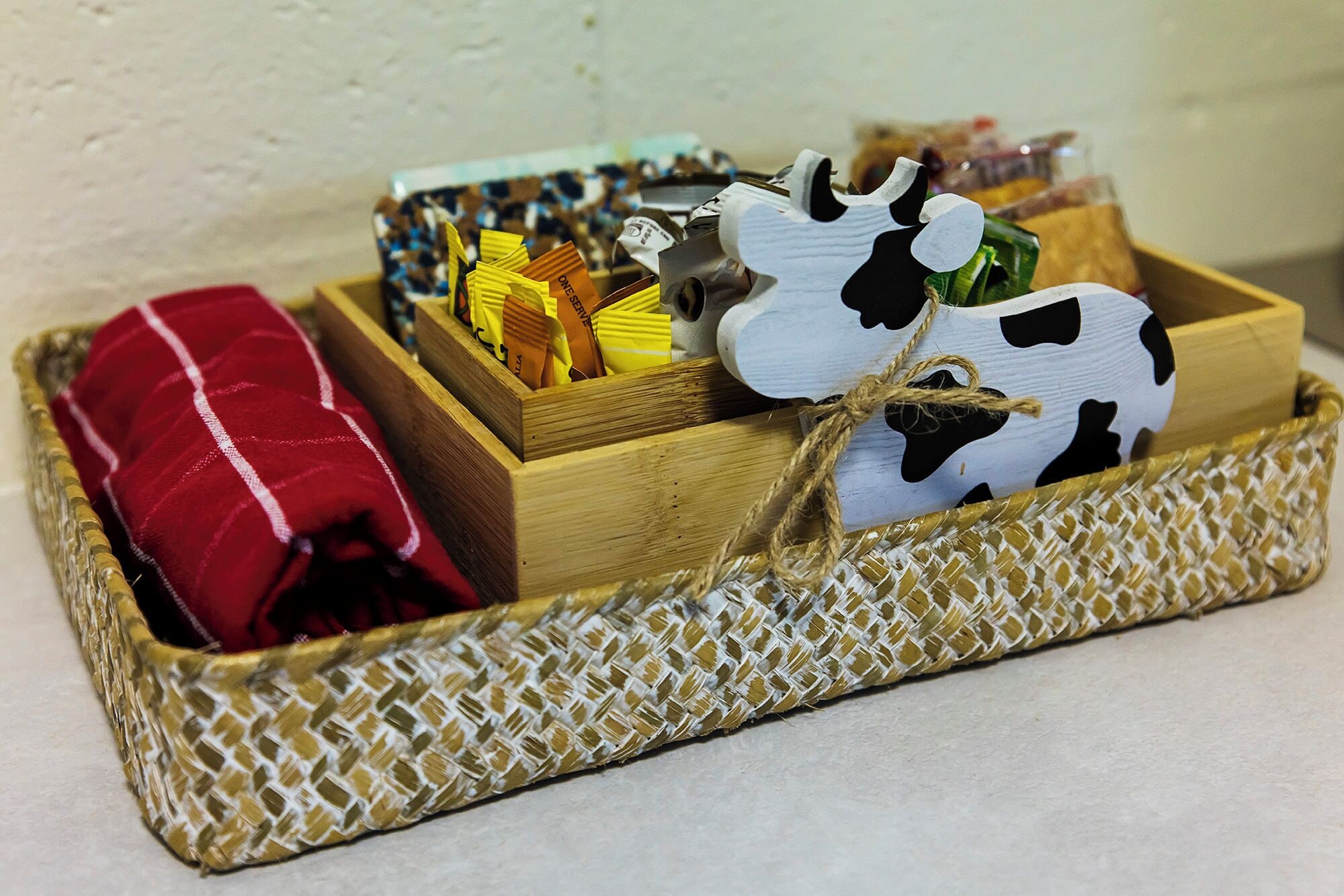 A wooden basket with a woven base containing a red pillow, a cow-shaped container, and a box with yellow items.