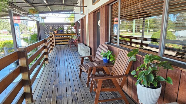 A wooden deck with a covered area, featuring a table and chairs, a potted plant, and a window with blinds. - Kilcoy Motel (Kilcoy)