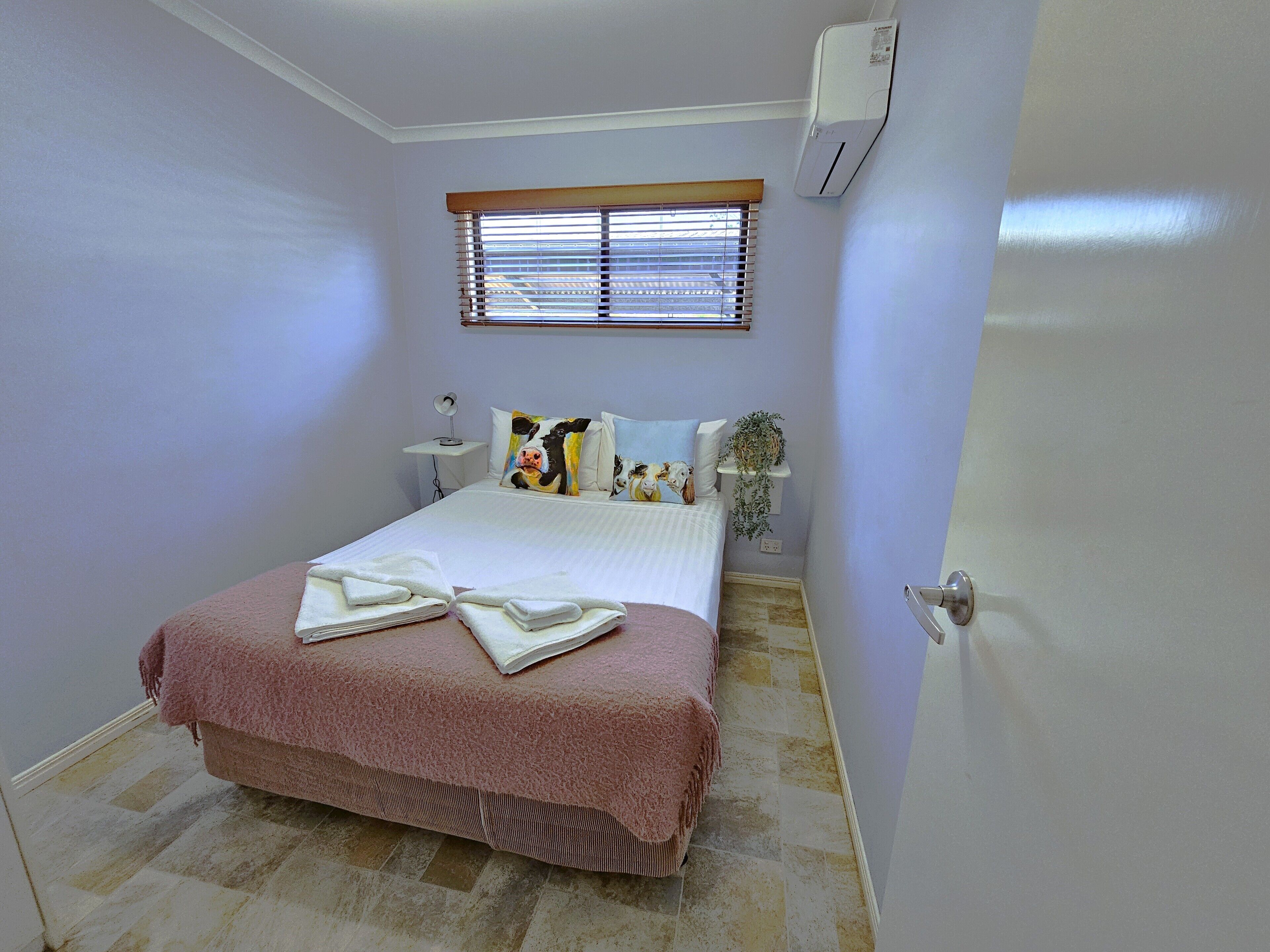A bedroom with a bed, a bedside table with a lamp, a window with blinds, and an air conditioning unit.