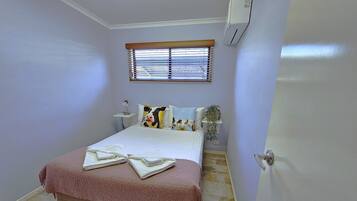 A bedroom with a bed, a bedside table with a lamp, a window with blinds, and an air conditioning unit.
