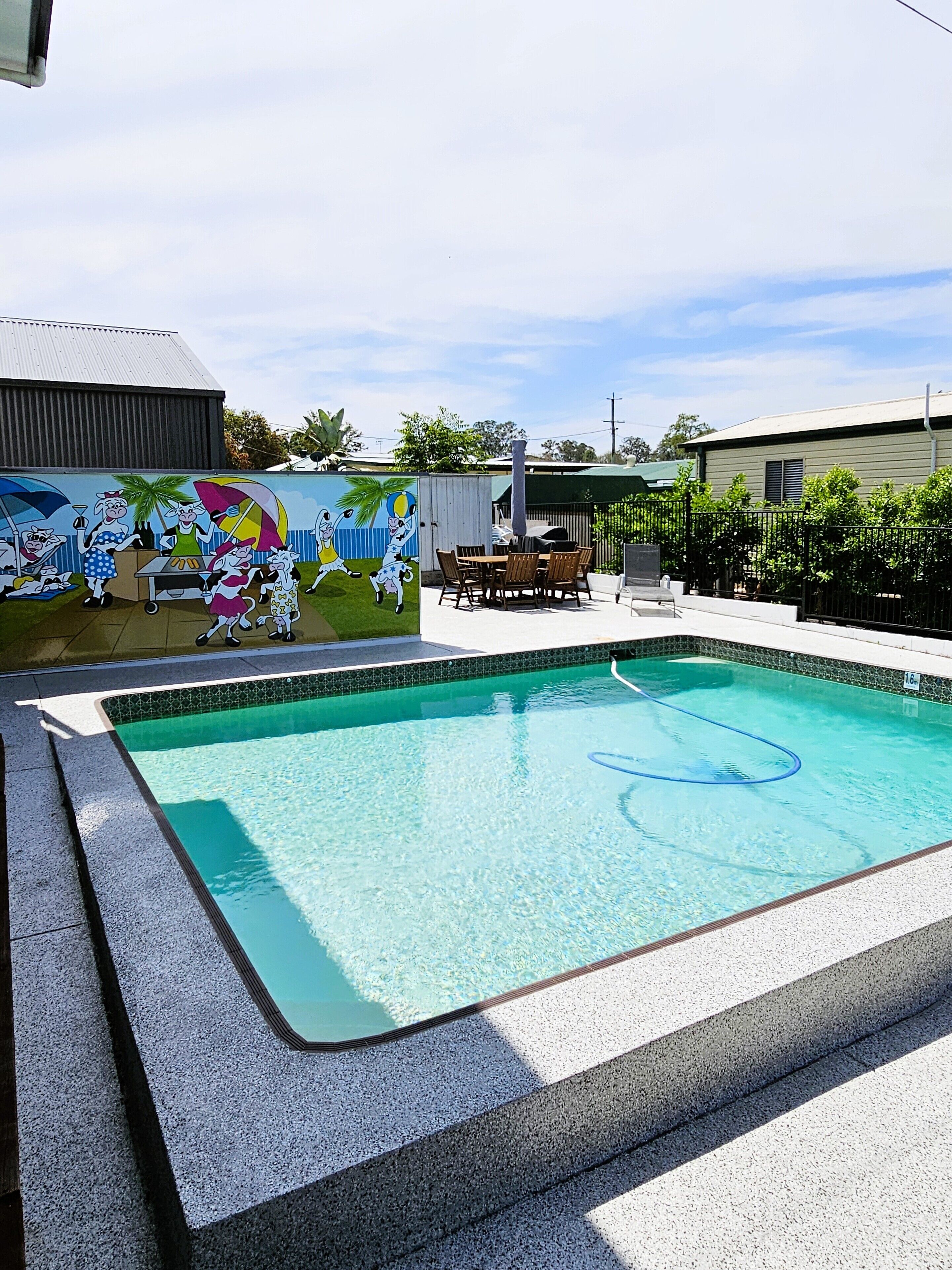 A swimming pool with a tiled edge, a mural on the wall, and a patio area with a table and chairs.