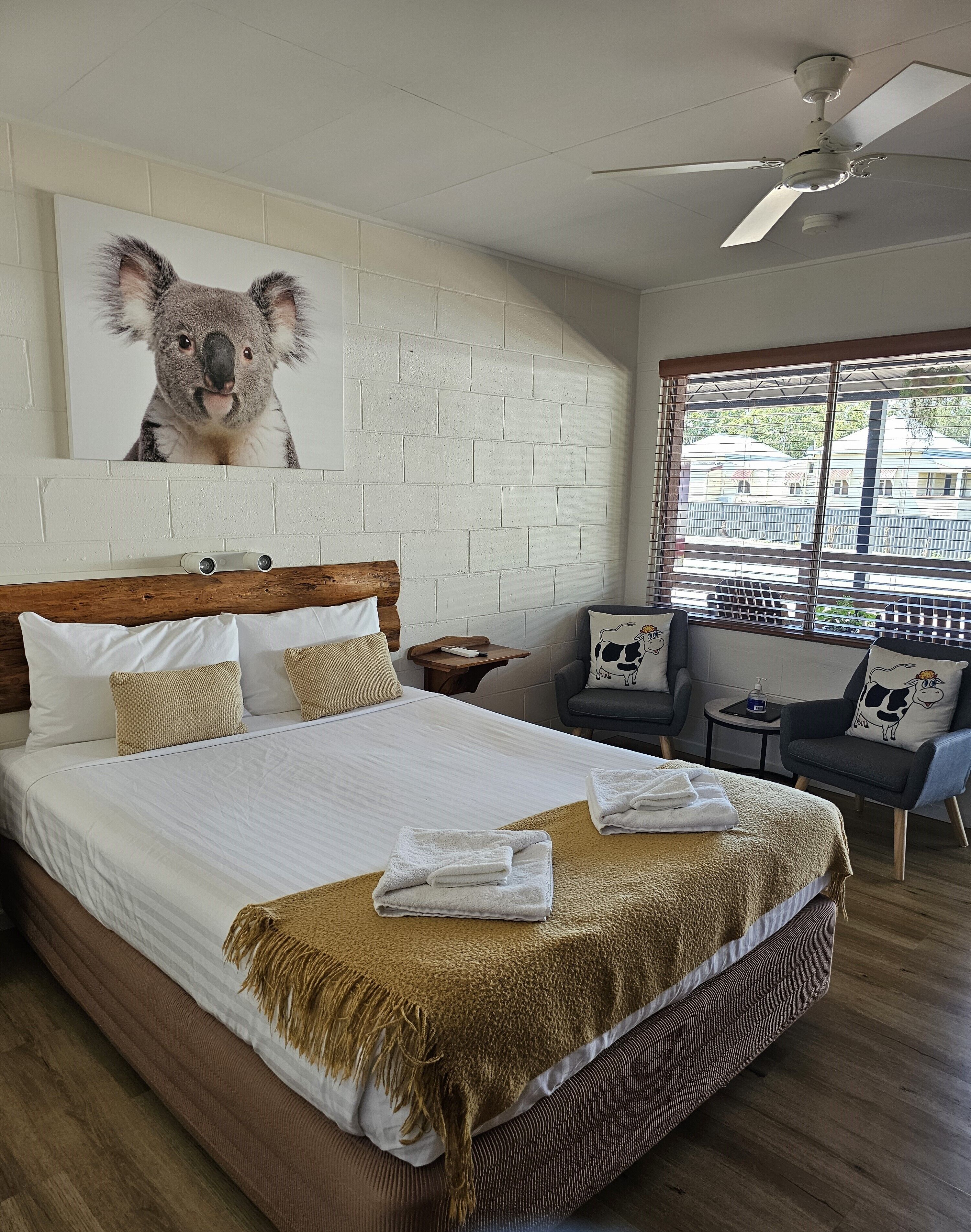 Een hotelkamer met een groot bed, een plafondventilator, een raam met uitzicht en een schilderij van een koala aan de muur.