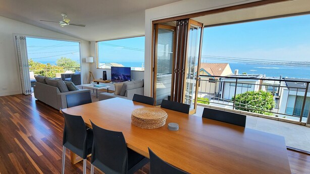 Un moderno salón con una mesa de comedor de madera, sillas negras, un sofá gris y un televisor. Amplias ventanas ofrecen vistas al mar y a edificios.