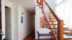 Un interior moderno con una escalera de madera, un pasillo con un banco y obras de arte en las paredes.