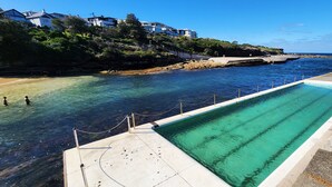 Una piscina junto a una costa rocosa con agua cristalina, un patio de hormigón y vistas a casas al fondo.