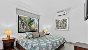 A bedroom with a bed, two bedside tables with lamps, a window with a view of trees, an air conditioner, and a framed artwork on the wall. - Amazing Condo! Playas del Coco (Provincia de Guanacaste)