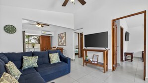 A living room with a blue sofa, a flat-screen TV on a wooden stand, a ceiling fan, and a dining area in the background. - Amazing Condo! Playas del Coco (Provincia de Guanacaste)