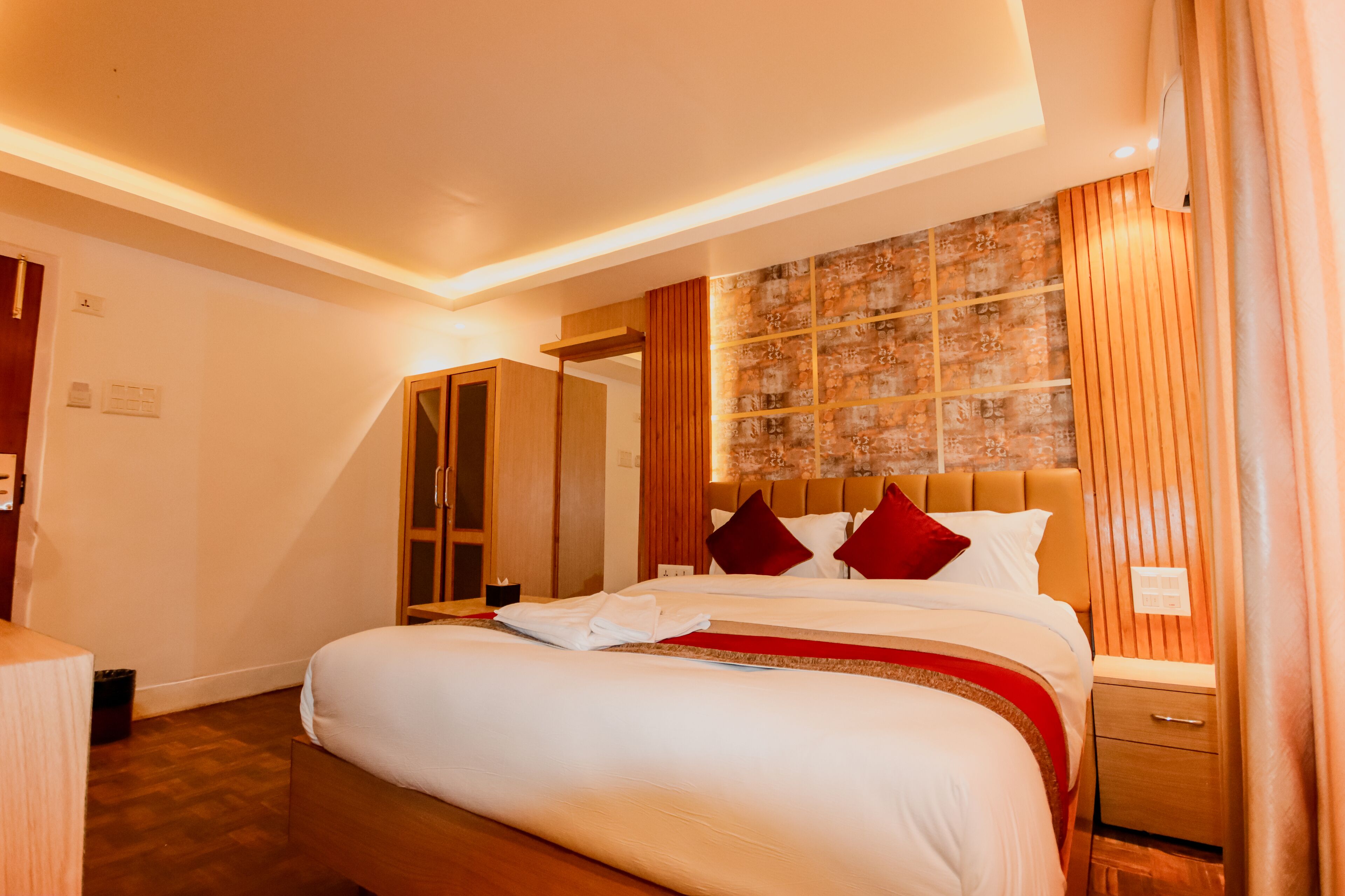 A hotel room with a large bed, wooden headboard, and a red accent pillow. There is a wooden nightstand and a small drawer.