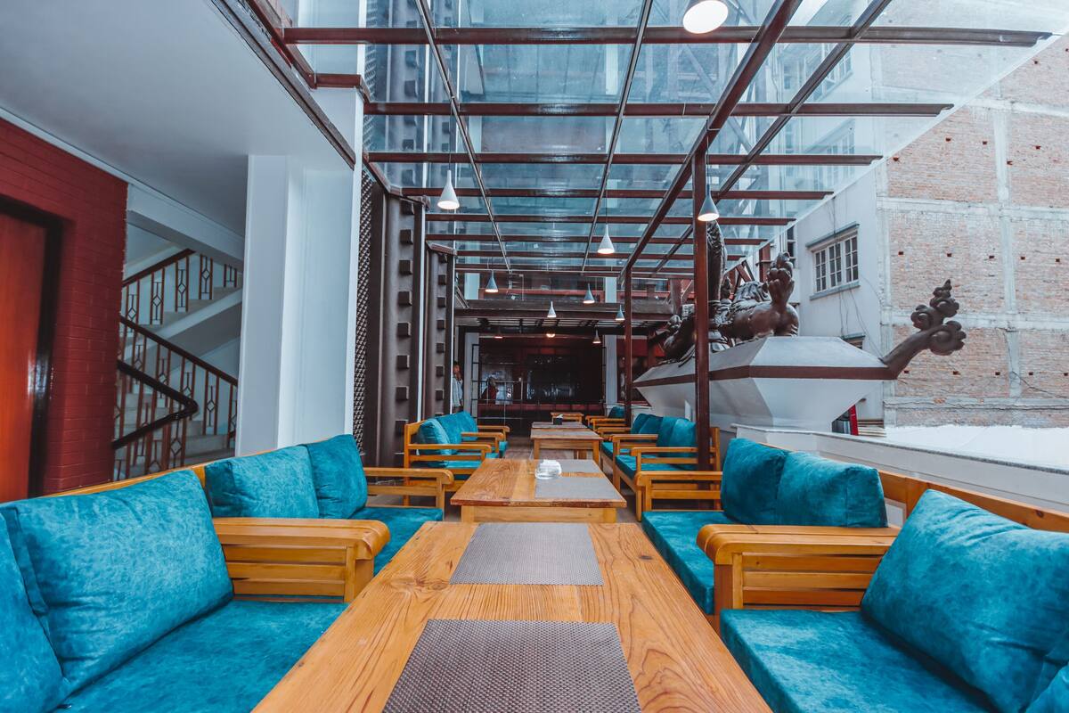 A modern interior with wooden tables and blue cushioned seating, a glass ceiling, and a brick wall.