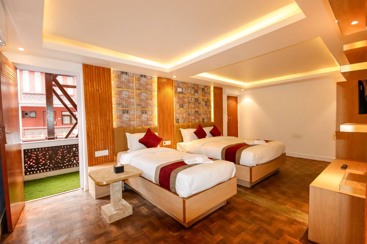 A hotel room with two beds, wooden flooring, a small table, and a view of a building outside.