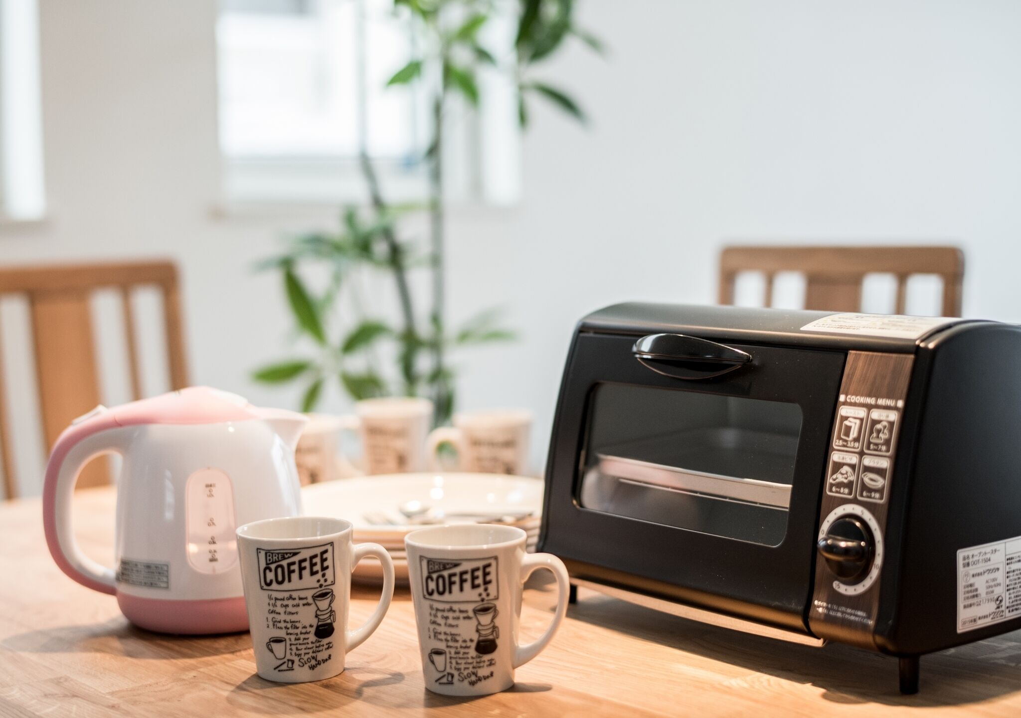 A kitchen with a black toaster oven, a pink kettle, and two coffee mugs on a wooden table.