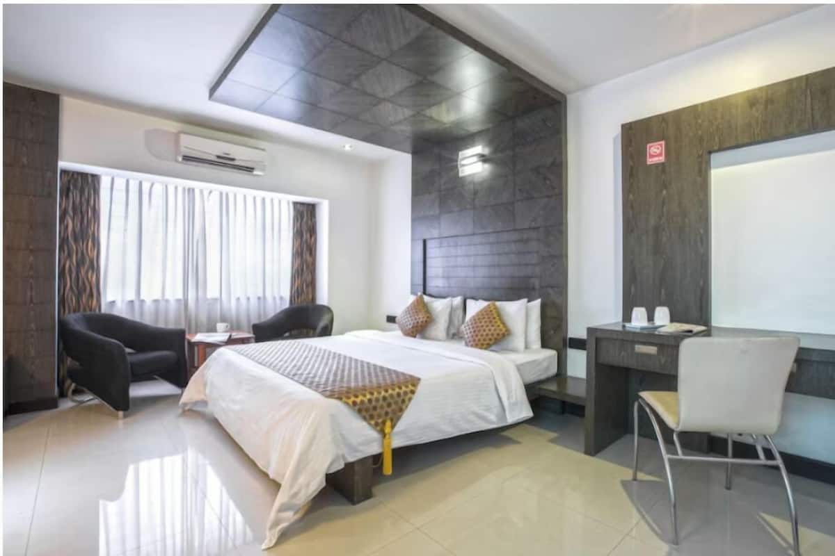 Habitación de hotel moderna con una cama grande, un escritorio y una silla. Se ve un equipo de aire acondicionado en la pared y una ventana con cortinas.