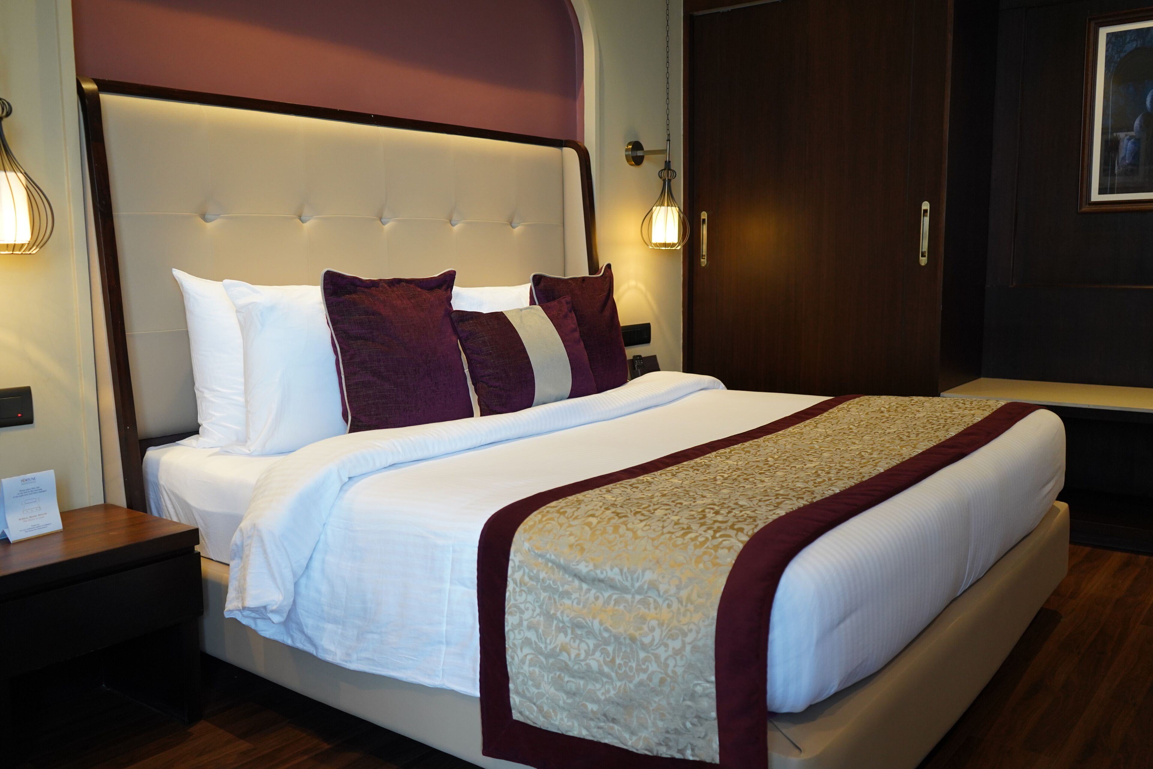 Cama bem arrumada com roupa de cama branca e bordô, cabeceira com luminária e mesa de cabeceira de madeira com abajur.