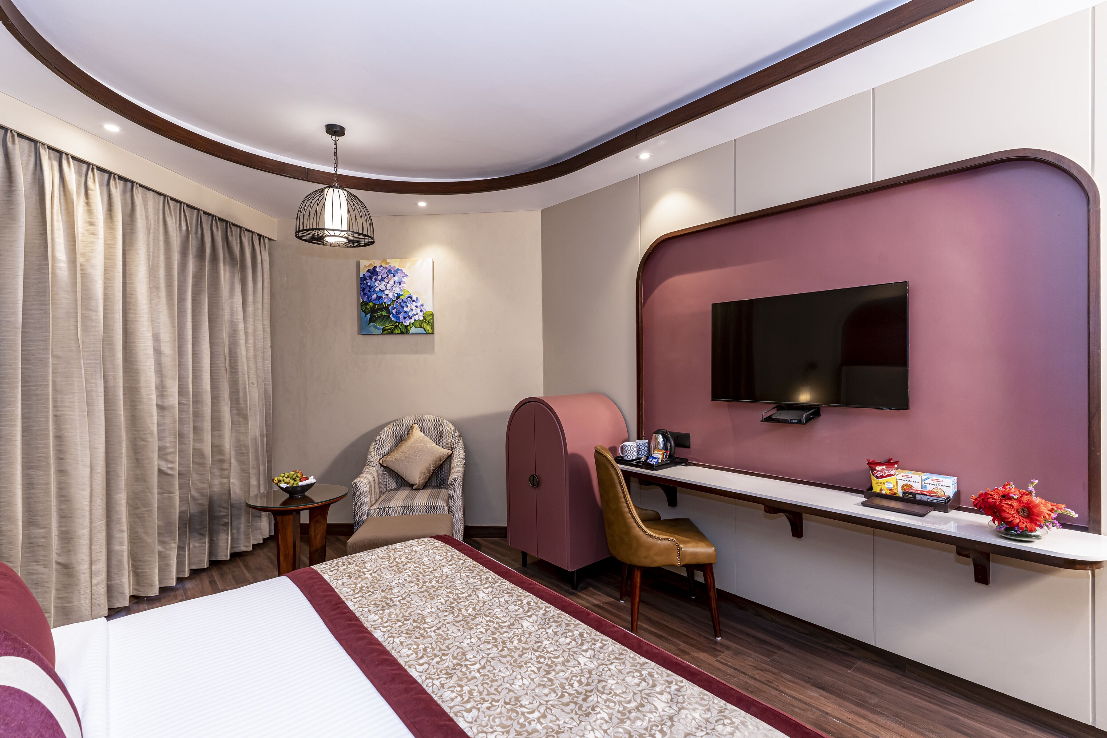 Ein modernes Hotelzimmer mit einem Bett, einem Schreibtisch mit Flachbildfernseher, einem Sessel, einem kleinen Tisch mit Obstschale und einer Wanddekoration.