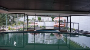 An indoor swimming pool with clear glass walls, a tiled floor, and a view of a building with a red roof and outdoor seating area. - Fortune Khajjiar - Member ITC Hotels' Group (Chamba)