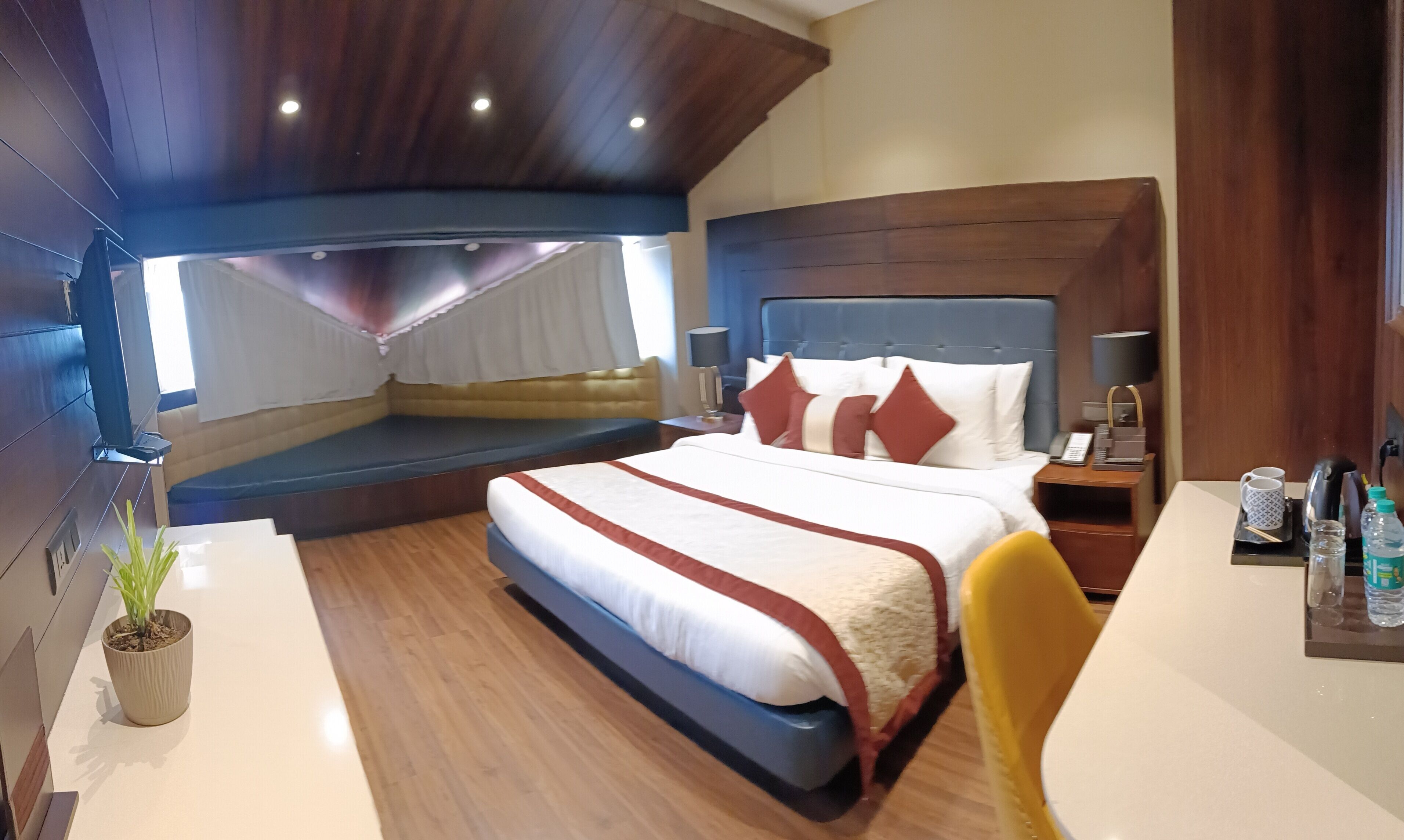 A modern hotel room with a large bed, a seating area, a desk, and a potted plant.