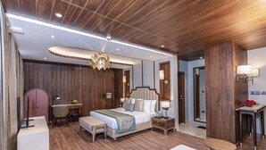A modern hotel room with a wooden ceiling, a large bed with a beige and white color scheme, a desk with a chair, a small table with a bowl of fruit, and a bathroom visible through an open door. - Fortune Khajjiar - Member ITC Hotels' Group (Chamba)