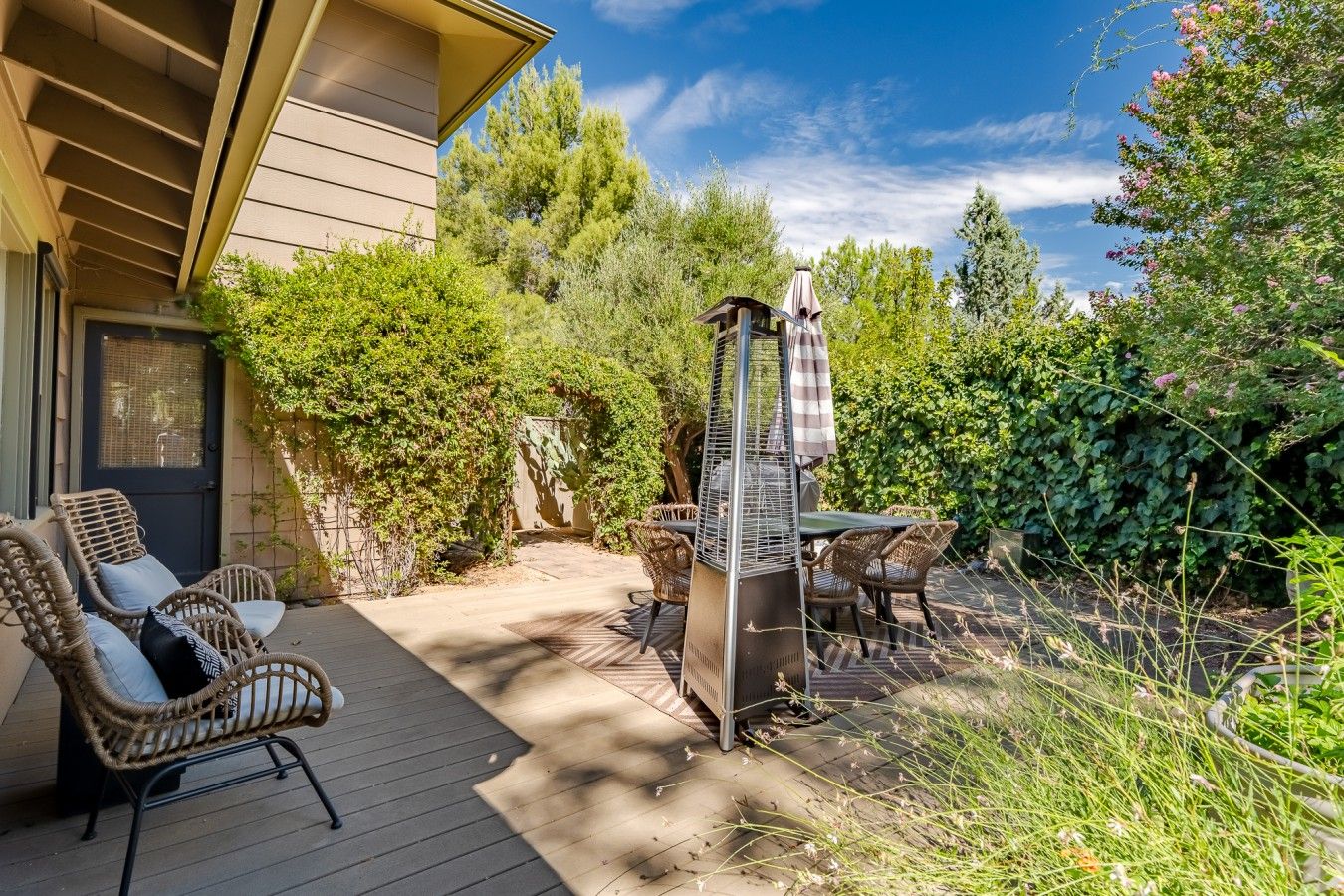 Une terrasse dotée de meubles en osier, d’un barbecue et offrant une vue sur une maison avec une porte et de la verdure.