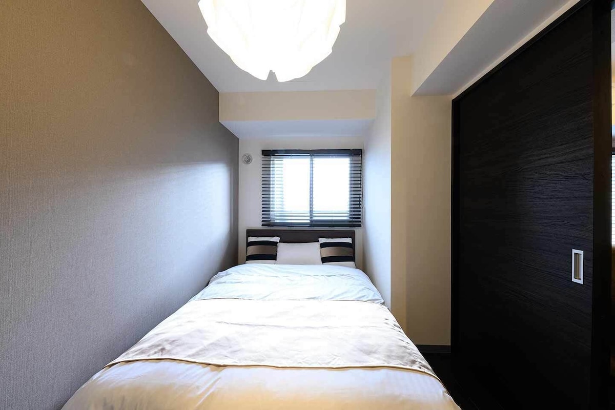 Une petite pièce avec un lit, une fenêtre munie de stores et une porte sombre.