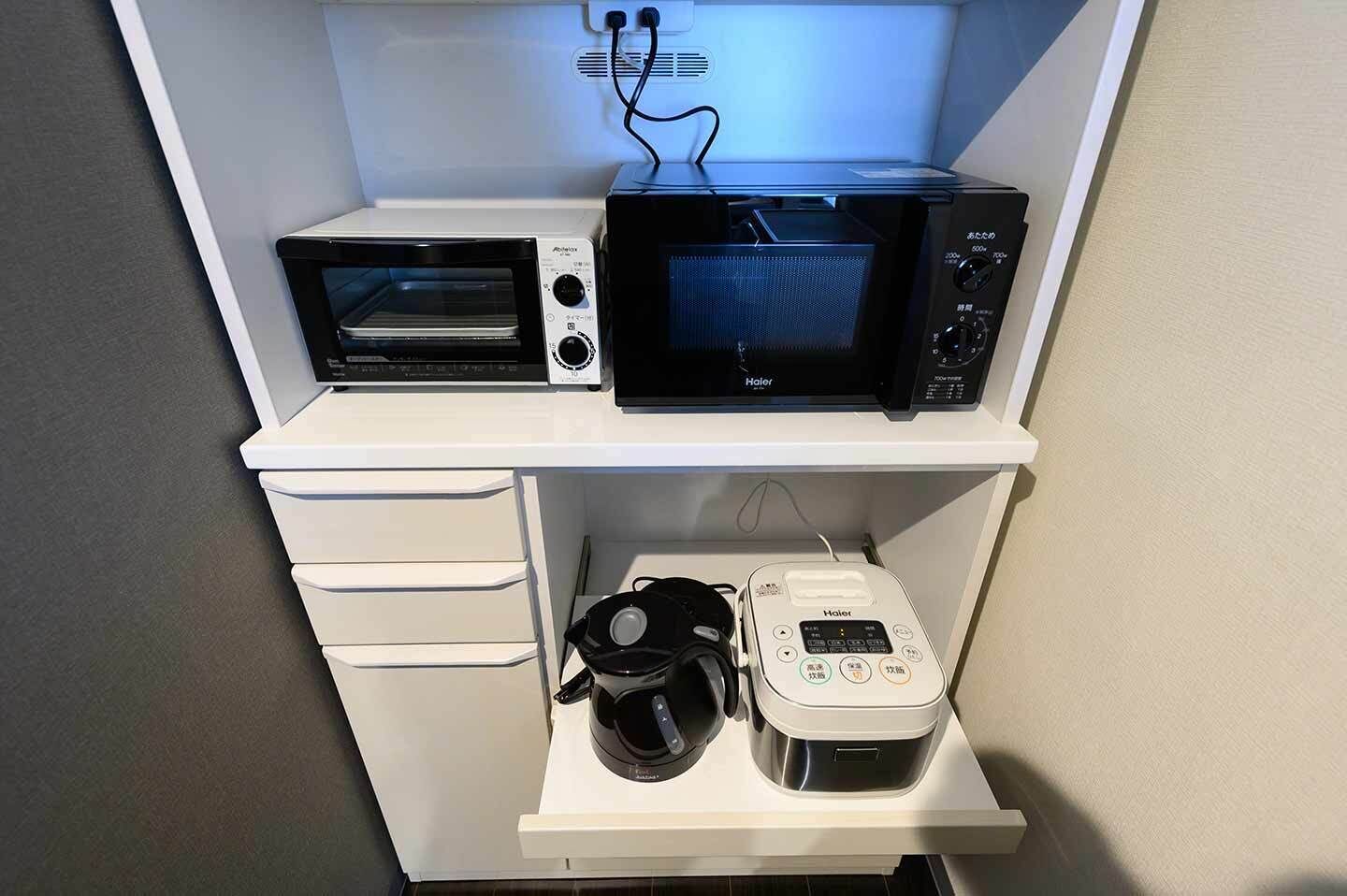 A compact kitchenette with a microwave, toaster, and coffee maker on a white shelf.