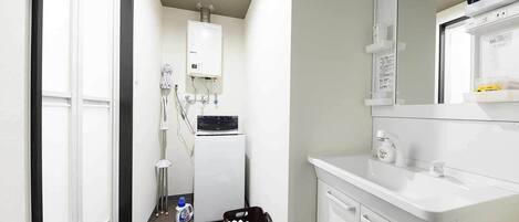 Un baño moderno con lavamanos blanco, espejo y toallero. Hay una zona de ducha con puerta de cristal y un inodoro blanco. Una maleta negra está en el suelo.