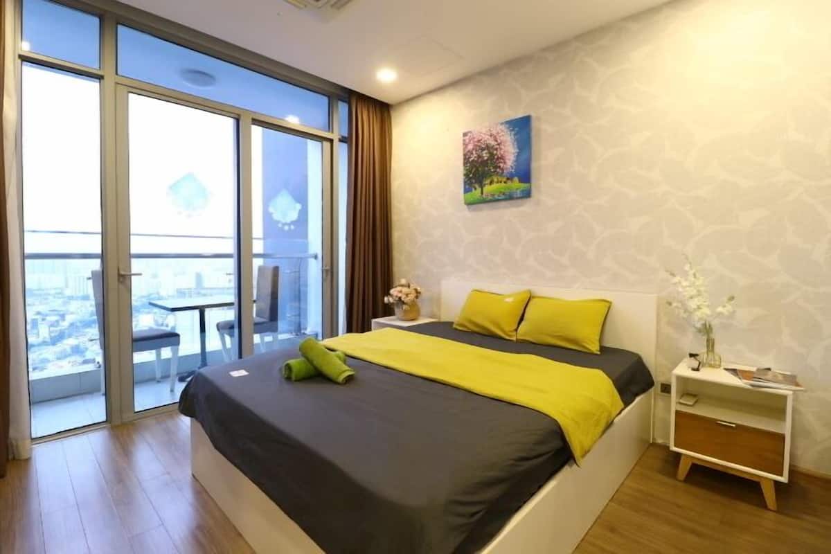 Une chambre d’hôtel moderne avec un grand lit, une table de chevet, une petite table et une vue sur la ville grâce à de grandes fenêtres.