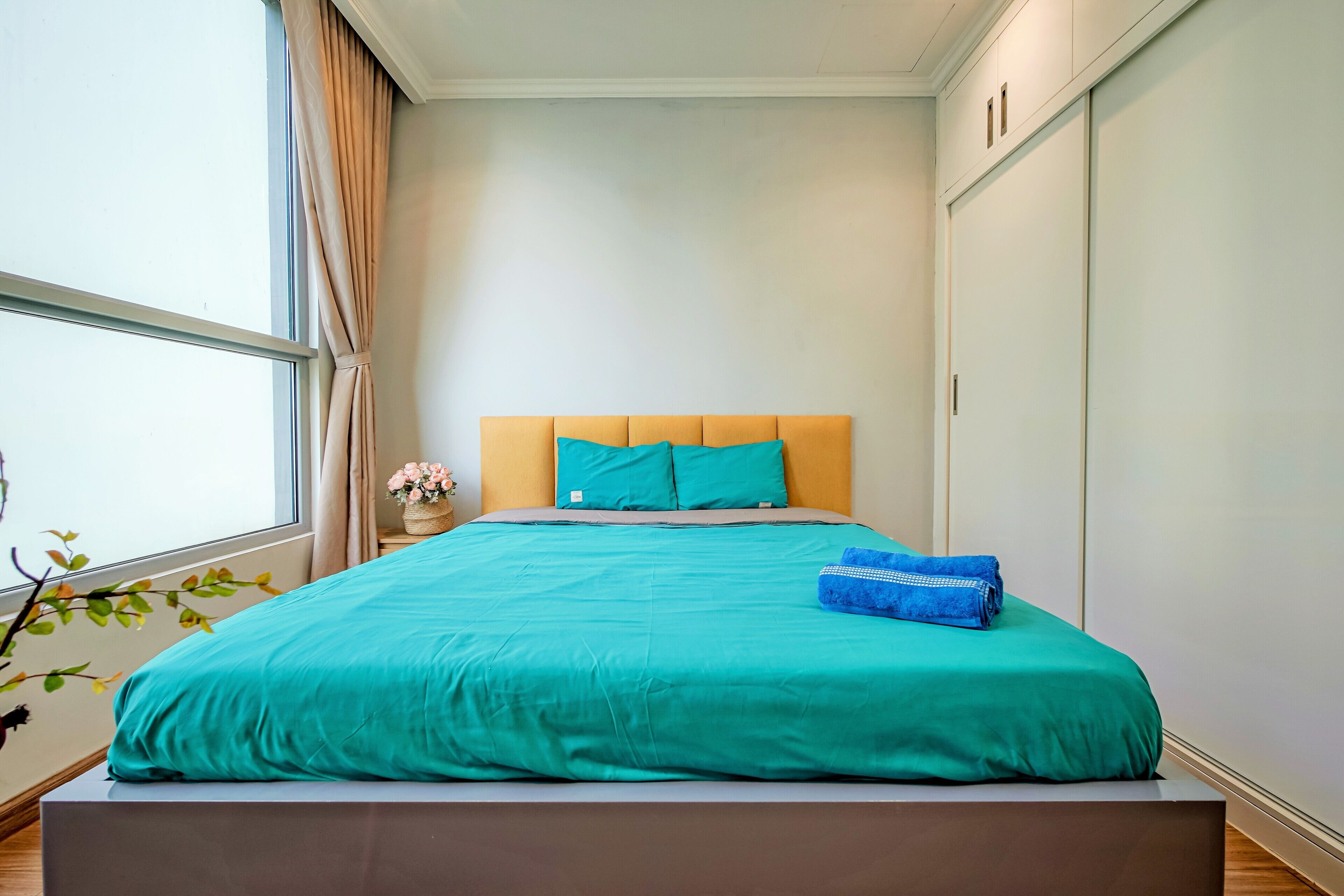 A neatly made bed with a teal bedspread and two pillows, a window with curtains, and a view of a tree outside.