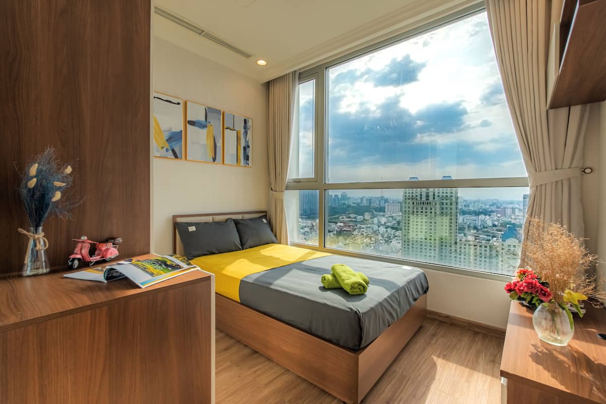 Une chambre moderne avec une grande fenêtre, un lit recouvert d’une couverture jaune, un bureau avec un magazine et un vase rempli de fleurs.