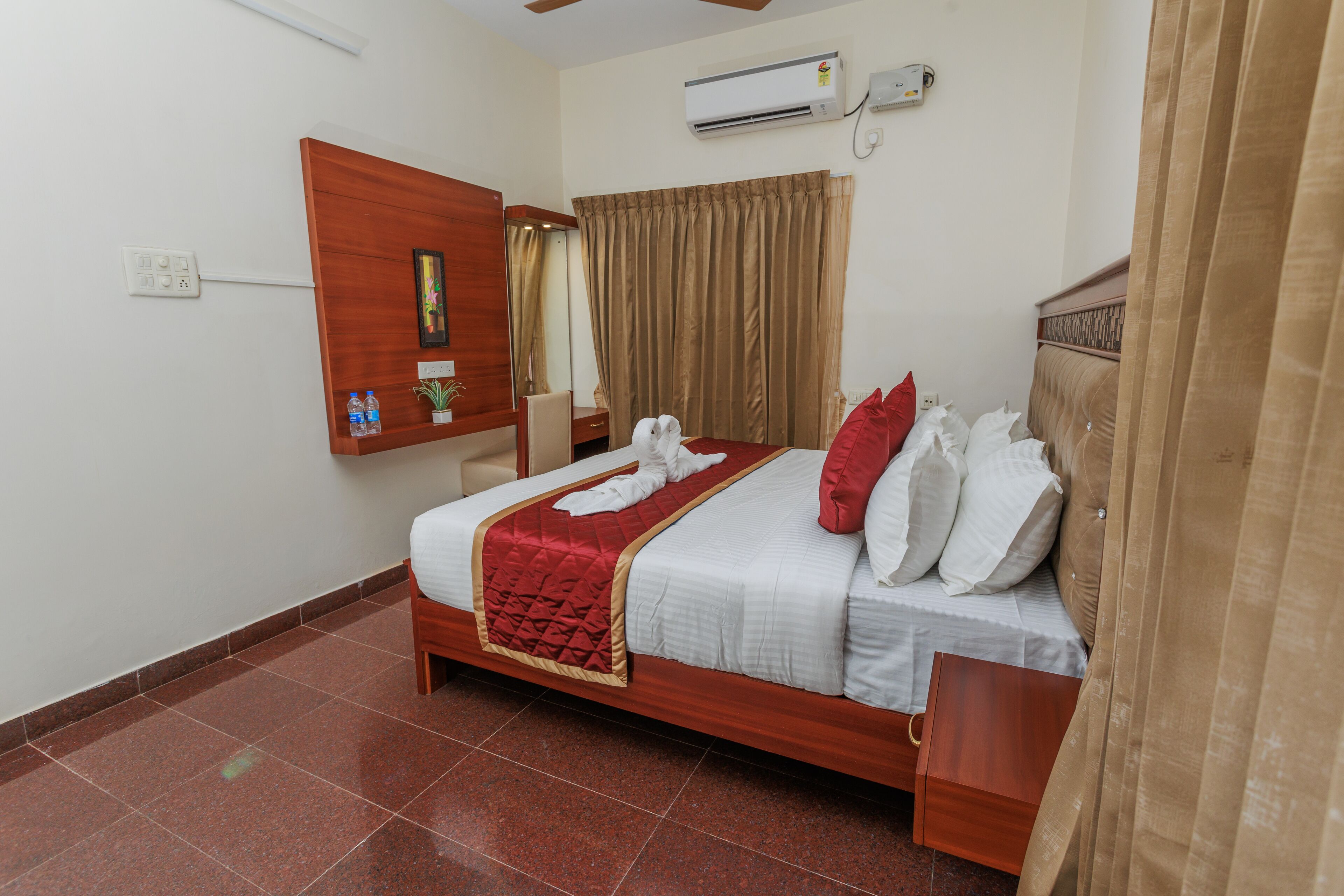 Habitación de hotel con cama, cabecera de madera, mesita de noche, espejo, aire acondicionado de pared y ventana con cortinas.