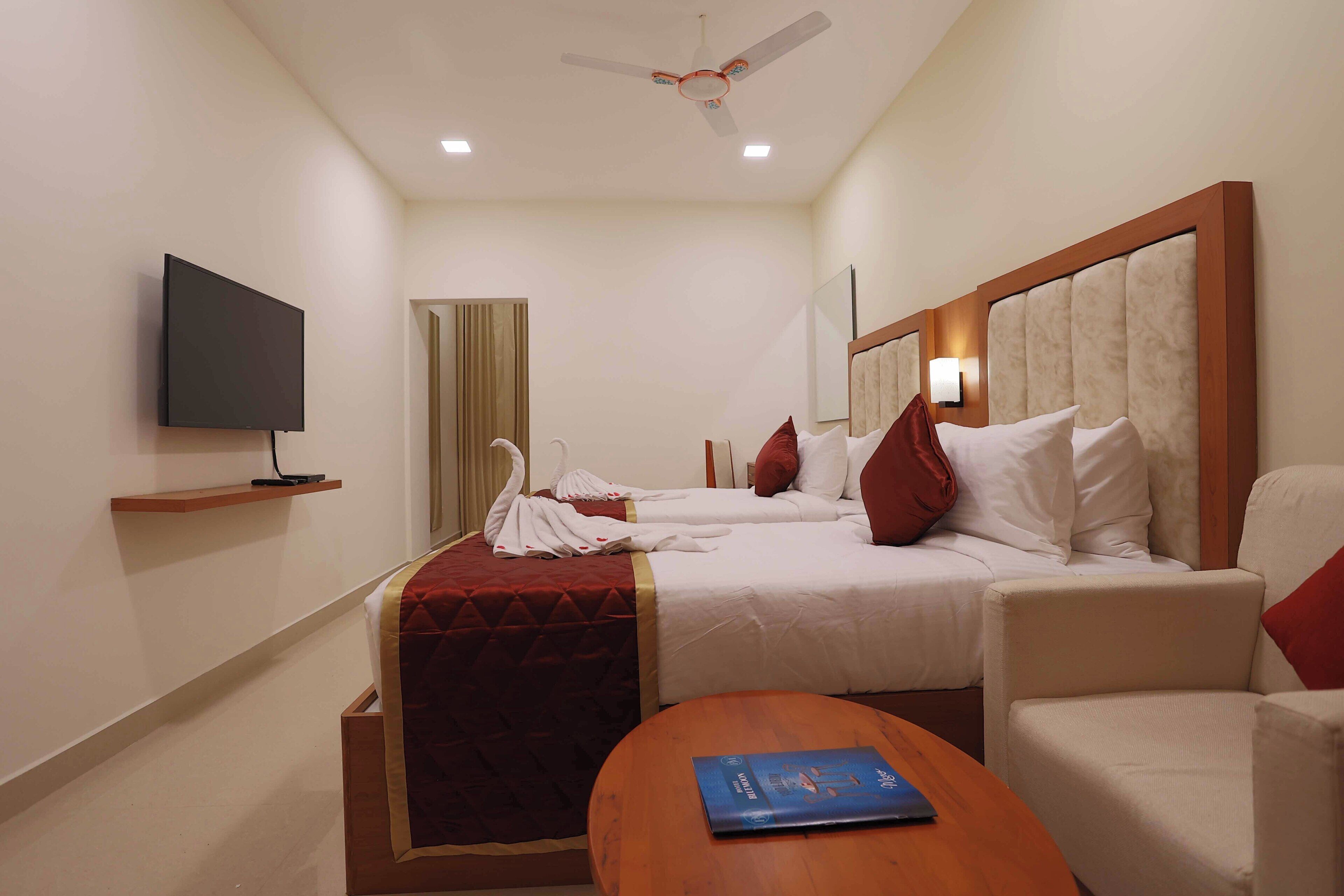 A hotel room with two beds, a TV, a ceiling fan, and a wooden table with a book.