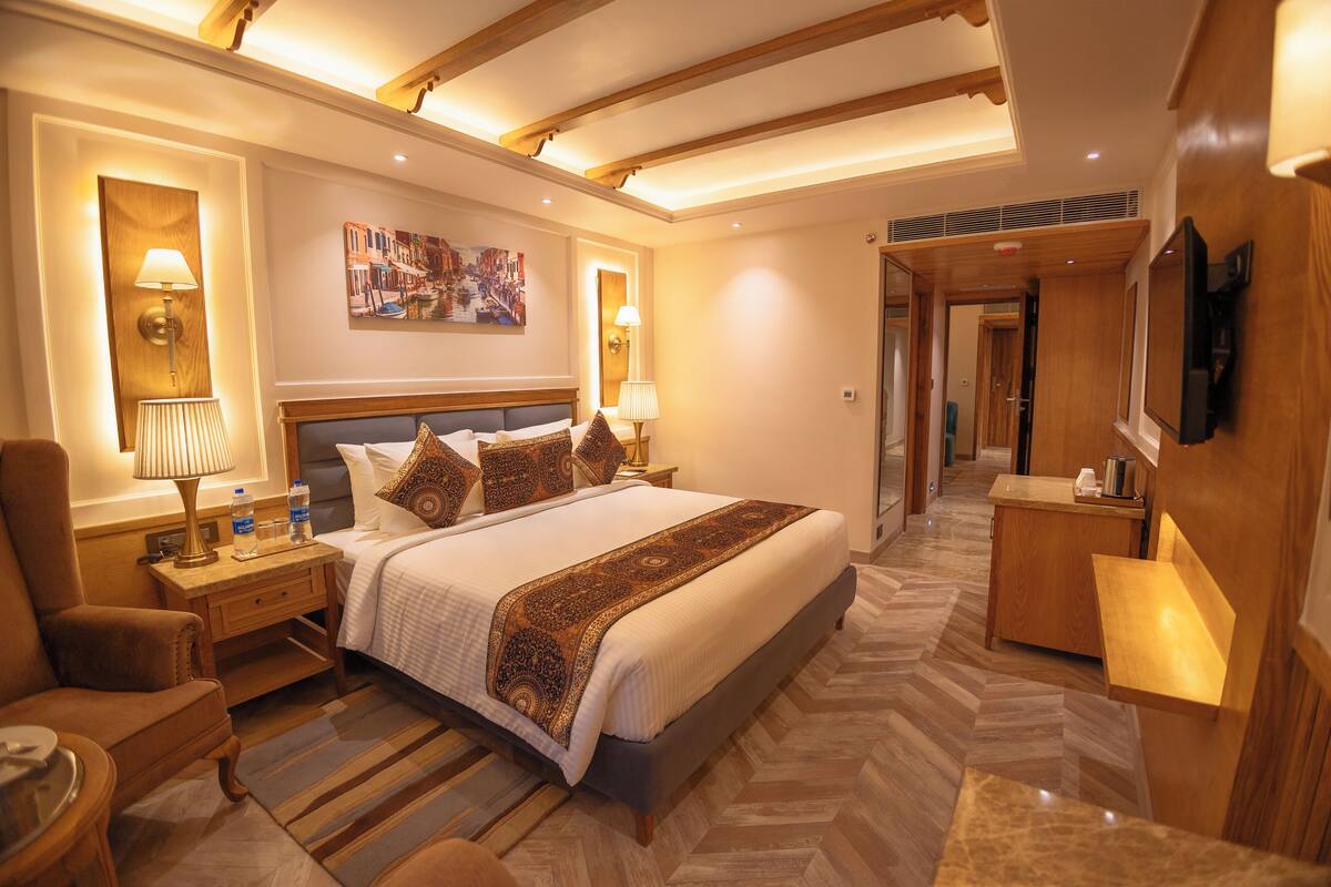 A spacious hotel room with a large bed, a sitting area with a chair, a desk, and a television.